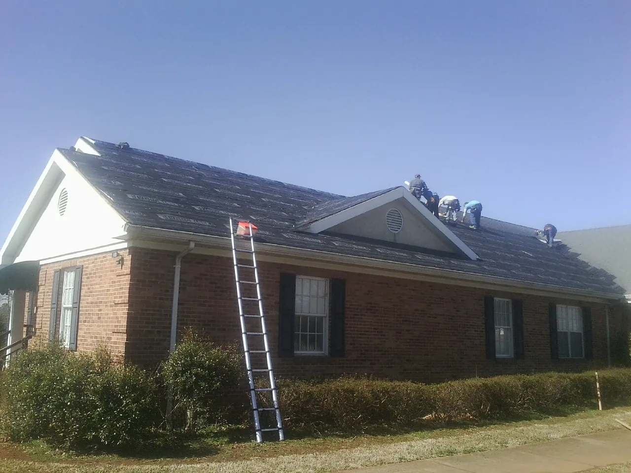 Crew tearing shingles off a brick ranch roof with a ladder set — Emergency Roof Repair work by our crew in Cuero