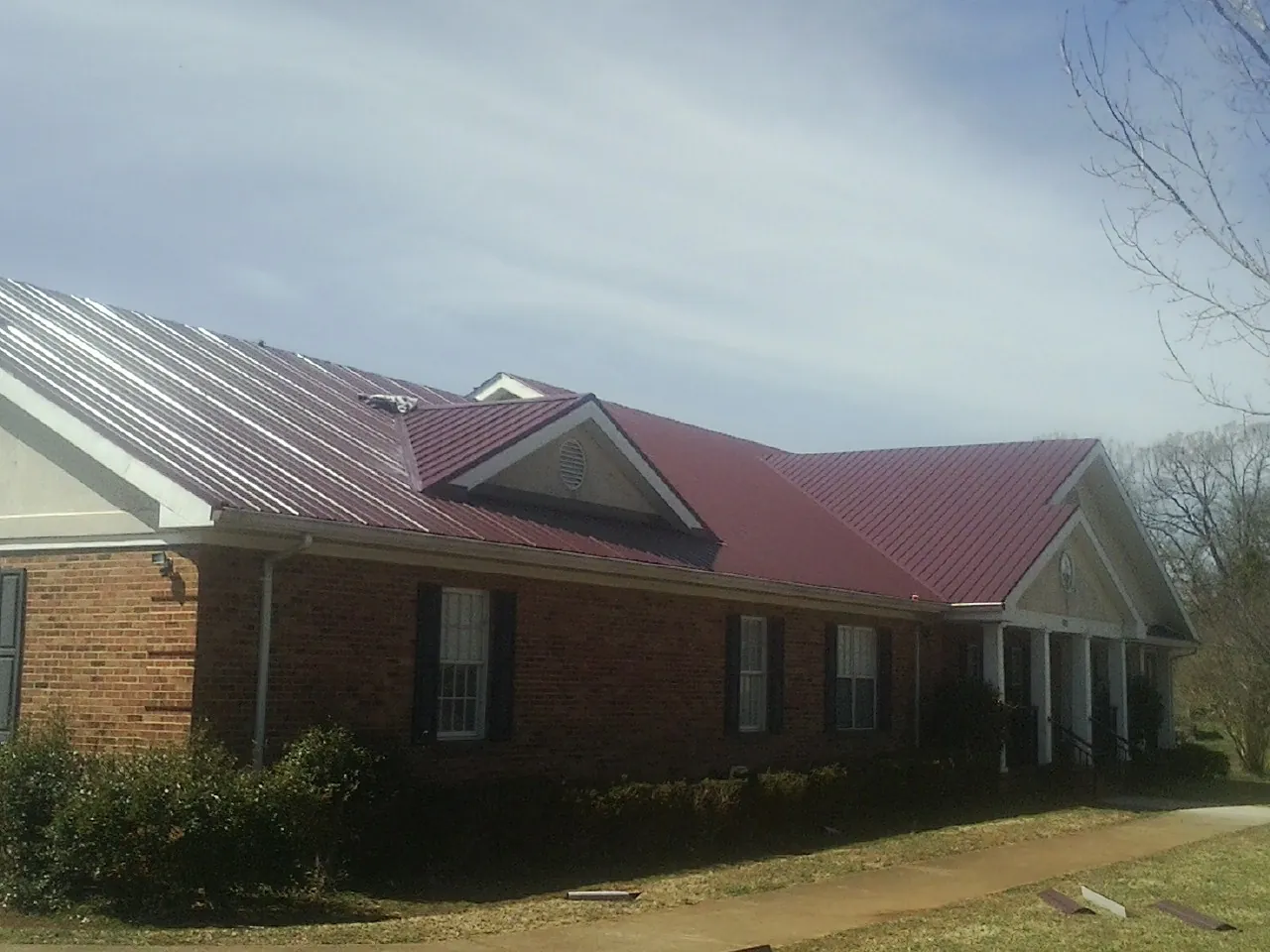 Red metal roof with gabled entries on a long brick building, from a Hail Damage Roof Repair job in Cuero