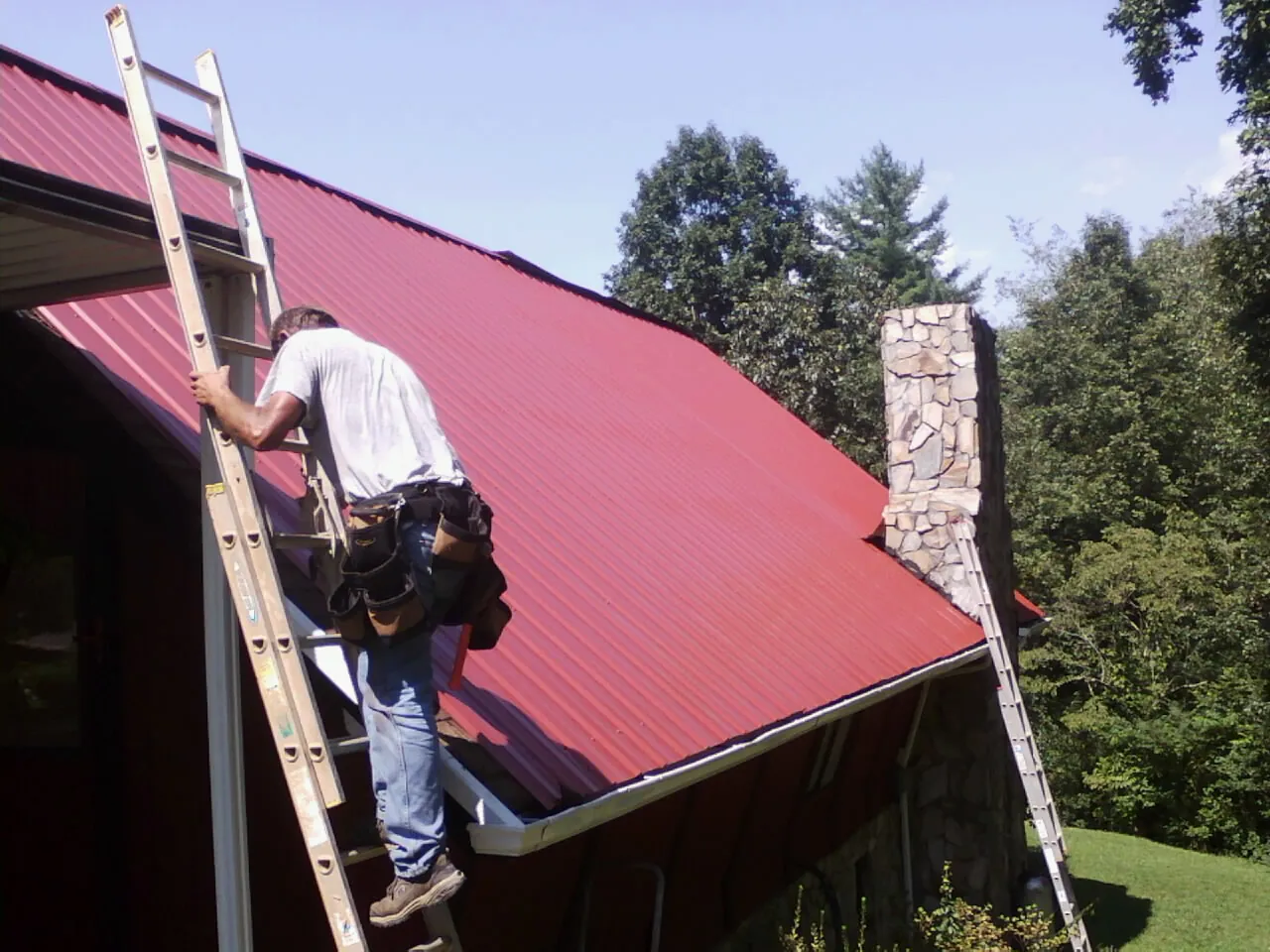 Roofer on a ladder beside a red metal roof and stone chimney — Soffit & Fascia Repair by Roofing Brothers in Cuero